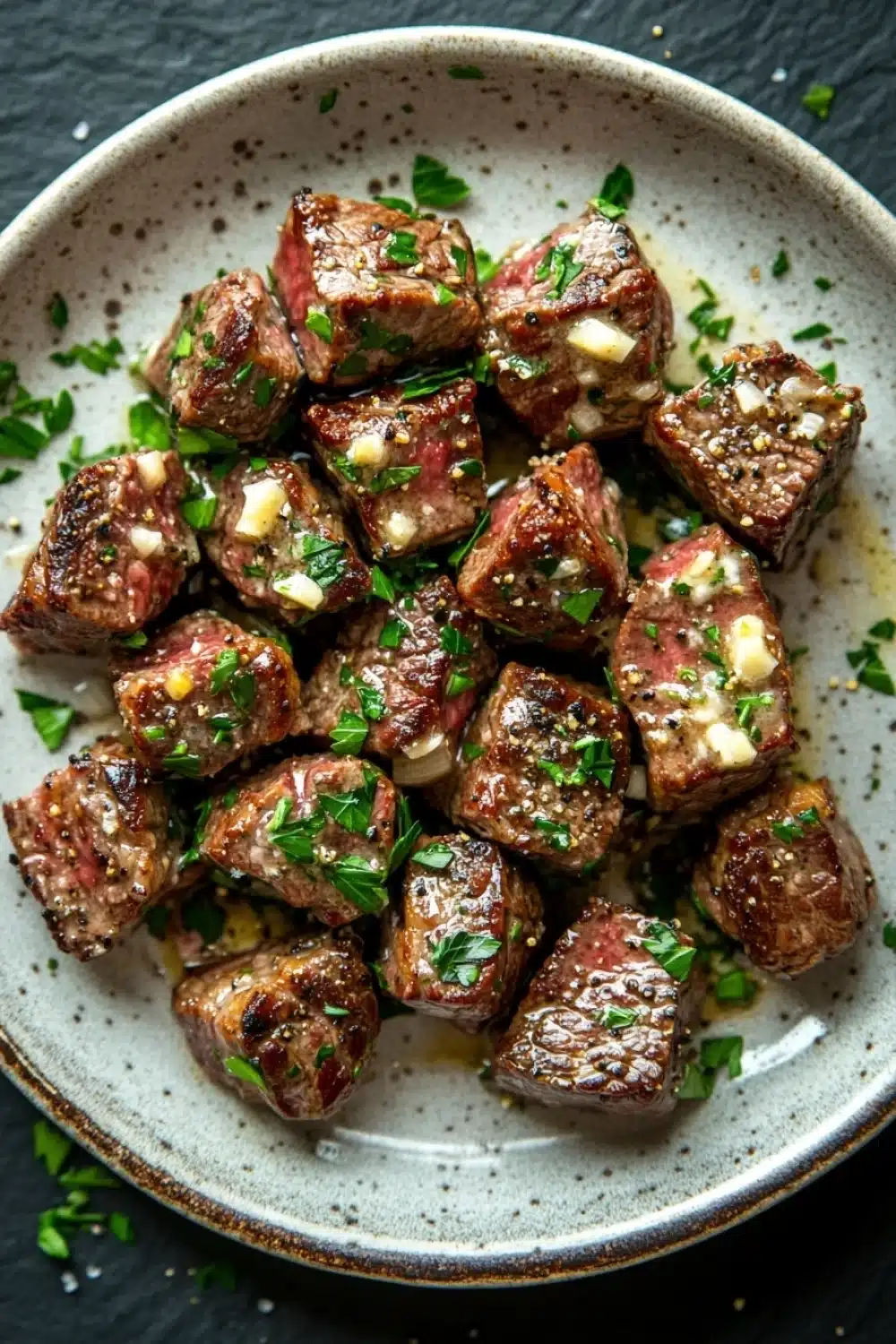 Steak bites with garlic butter stove top - the image is a close-up of a plate of food. the plate is round and has a light blue and white speckled pattern. on top of the plate, there are several pieces of cooked meat, which appear to be steak or beef tenderloin. the meat is garnished with chopped herbs and a dollop of white cheese. the dish is served on a dark grey countertop.