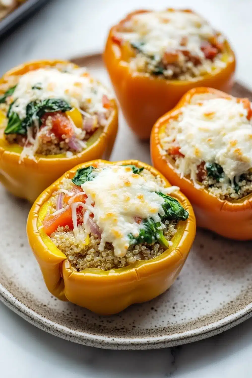 Stuffed bell peppers ricotta - the image shows four stuffed bell peppers on a white plate. the bell peppers are yellow in color and have a crispy texture. they are filled with a mixture of quinoa, tomato, and spinach, and topped with melted cheese. the plate is sitting on a marble countertop and there is a baking tray in the background.