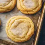 Sugar cookie chewy - the image shows a baking tray with six freshly baked cookies on it. the cookies are round and golden brown in color, with a criss-cross pattern on top. they are dusted with powdered sugar, giving them a shiny appearance. the tray is lined with parchment paper, and the cookies are arranged in a row on the tray. the background is a dark wooden table.