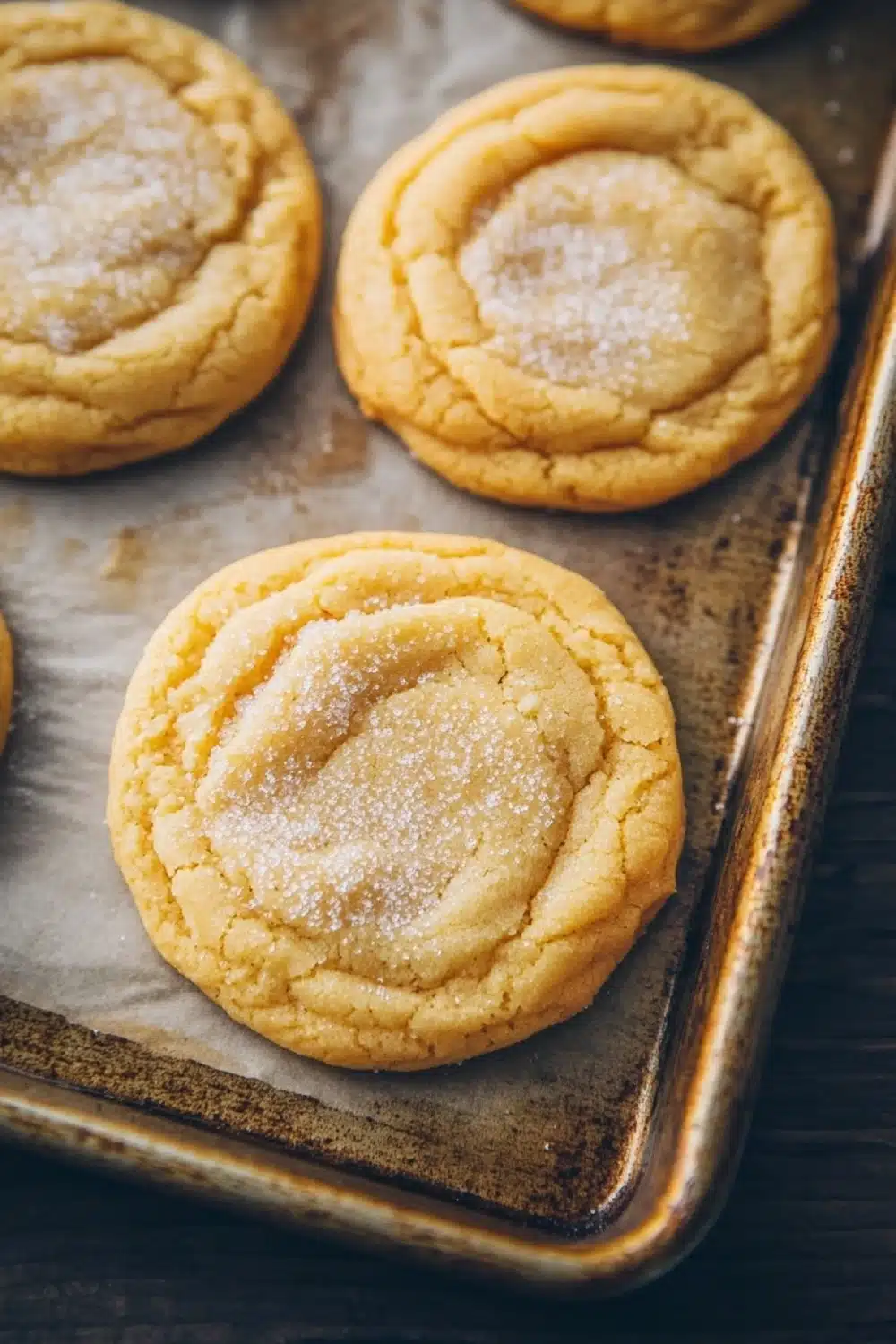 Sugar cookie chewy - the image shows a baking tray with six freshly baked cookies on it. the cookies are round and golden brown in color, with a criss-cross pattern on top. they are dusted with powdered sugar, giving them a shiny appearance. the tray is lined with parchment paper, and the cookies are arranged in a row on the tray. the background is a dark wooden table.
