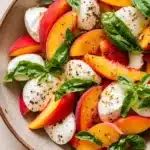 Summer peach caprese salad - the image is a close-up of a salad in a bowl. the salad is made up of sliced peaches, mozzarella cheese, and fresh basil leaves. the peaches are arranged in a circular pattern, with some overlapping each other. the cheese is white and fluffy, and the basil leaves are a vibrant green color. the bowl is a light brown color and is sitting on a wooden table. the overall presentation of the salad is fresh and appetizing.