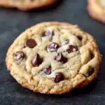 Thin and crispy chocolate chip cookie - the image shows a close-up of a freshly baked chocolate chip cookie on a black surface. the cookie is round and golden brown in color, with chocolate chips scattered throughout. it appears to be soft and chewy, with a slightly crumbly texture. in the background, there are more chocolate chip cookies scattered around the surface. a knife is resting on the right side of the image, suggesting that the cookie is being prepared for baking.