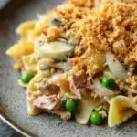 Tuna casserole no cheese - the image shows a plate of pasta dish on a dark grey plate. the pasta appears to be fettuccine alfredo, with a layer of breadcrumbs on top. the dish is garnished with sliced onions, green peas, and chunks of ham. the plate is sitting on a wooden table with a blurred background.