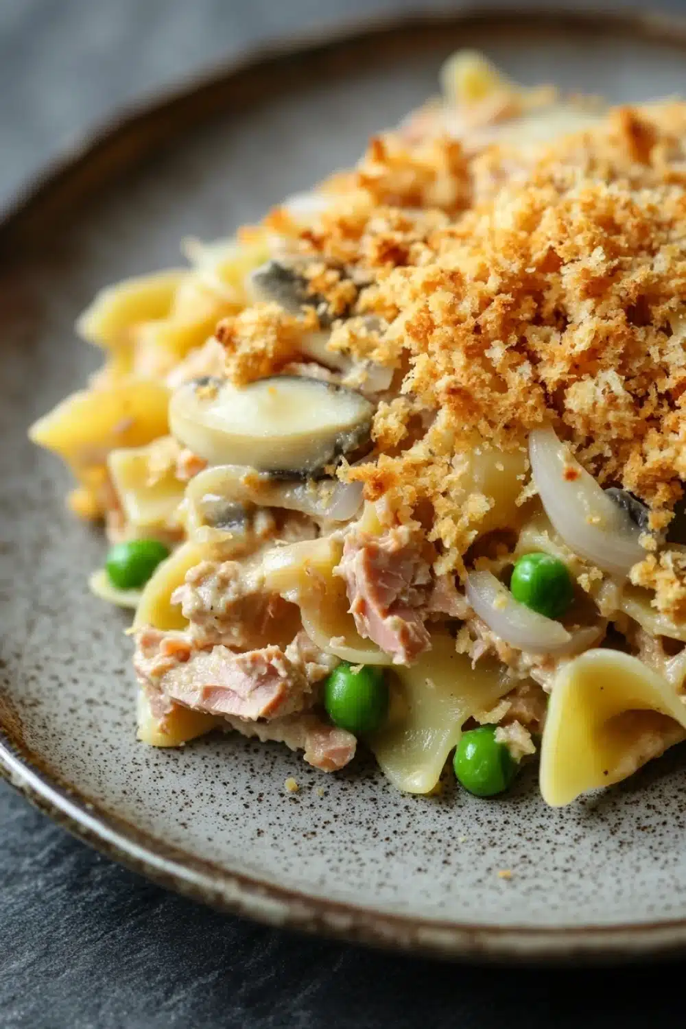 Tuna casserole no cheese - the image shows a plate of pasta dish on a dark grey plate. the pasta appears to be fettuccine alfredo, with a layer of breadcrumbs on top. the dish is garnished with sliced onions, green peas, and chunks of ham. the plate is sitting on a wooden table with a blurred background.