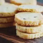 Vanilla bean shortbread cookie - the image shows a stack of freshly baked cookies on a wooden cutting board. the cookies are golden brown in color and have a crumbly texture. they are arranged in a neat stack, with some overlapping each other. the background is blurred, but it appears to be a dark countertop. the overall mood of the image is warm and inviting.