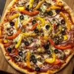 Vegan sheet pan pizza - the image is of a large pizza on a wooden cutting board. the pizza is round and has a golden brown crust. it is topped with a variety of toppings, including sliced red and yellow bell peppers, sliced mushrooms, red onions, and black olives. the toppings are arranged in a circular pattern, with some overlapping each other. the cutting board is placed on a dark wooden table, and there are two red bell peppers on the right side of the image.