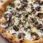 White sauce truffle pizza - the image is of a freshly baked pizza on a wooden cutting board. the pizza has a golden brown crust and is topped with melted cheese and sliced mushrooms. the mushrooms are arranged in a circular pattern on top of the cheese, with some of them overlapping each other. there are also small sprigs of fresh herbs scattered throughout the pizza. the background is blurred, but it appears to be a kitchen countertop.