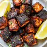 Air fryer salmon bites blackened - the image shows a plate of grilled salmon cubes. the salmon cubes are golden brown and appear to be seasoned with herbs and spices. they are arranged in a circular pattern on the plate, with two lemon wedges on either side. the plate is white and has a textured surface. the background is blurred, but it appears to be a kitchen countertop.