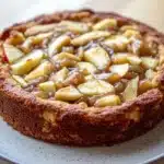 Apple cake with apple pie filling - the image is of a freshly baked apple tart on a white plate. the tart has a golden brown crust and is topped with sliced apples arranged in a circular pattern. the apples are cut into small cubes and are drizzled with a caramel sauce. the plate is sitting on a wooden table with a blurred background.