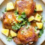 Baked chicken thighs pineapple - the image shows a plate of food with two pieces of grilled chicken on it. the chicken is golden brown and appears to be seasoned with herbs and spices. the plate is light blue with a speckled pattern and is placed on a wooden table. there are also slices of pineapple and chopped green onions scattered around the plate. the dish looks appetizing and ready to eat.