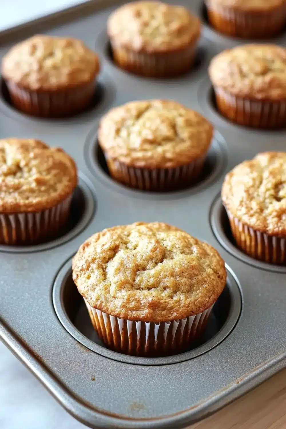 Banana muffin pictures - the image shows a muffin tin filled with freshly baked muffins. the muffins are golden brown in color and have a crumbly texture on top. they are arranged in a circular pattern in the tin, with each muffin slightly overlapping the one below it. the tin is sitting on a wooden countertop, and the background is blurred, but it appears to be a kitchen countertop.