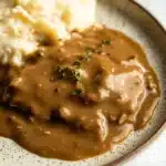 Beef chicken gravy - the image shows a plate of food with a serving of mashed potatoes and gravy. the plate is white with a brown rim and has a speckled pattern around the edges. the gravy is a rich, dark brown color and appears to be thick and creamy. on top of the gravy, there is a small sprig of thyme, adding a pop of green color to the dish. the mashed potatoes are white and fluffy, and the gravy is thick and thick. the dish is served on a white marble countertop.