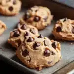 Chewy cookie dough - the image shows a baking tray lined with freshly baked chocolate chip cookies. the cookies are golden brown and appear to be soft and chewy. they are arranged in a single layer on the tray, with some overlapping each other. the tray is placed on a wooden table, and the background is blurred, making the cookies the focal point of the image.