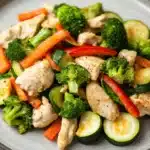 Chicken and veggie freezer packs - the image is a close-up of a plate of stir-fry with chicken, broccoli, zucchini, and carrots. the chicken is cut into small pieces and is arranged in a circular pattern on the plate. the vegetables are bright green and appear to be seasoned with herbs and spices. the plate is gray and has a textured surface. the background is blurred, but it appears to be a kitchen countertop.