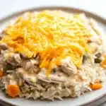 Chicken rice casserole crockpot - the image shows a plate of food with a serving of rice and chicken. the rice is white and fluffy, and the chicken is covered in a creamy sauce. on top of the rice, there is a layer of shredded cheese, which is a bright yellow color. the dish is garnished with small orange carrots and green bell peppers. the plate is sitting on a white marble countertop.