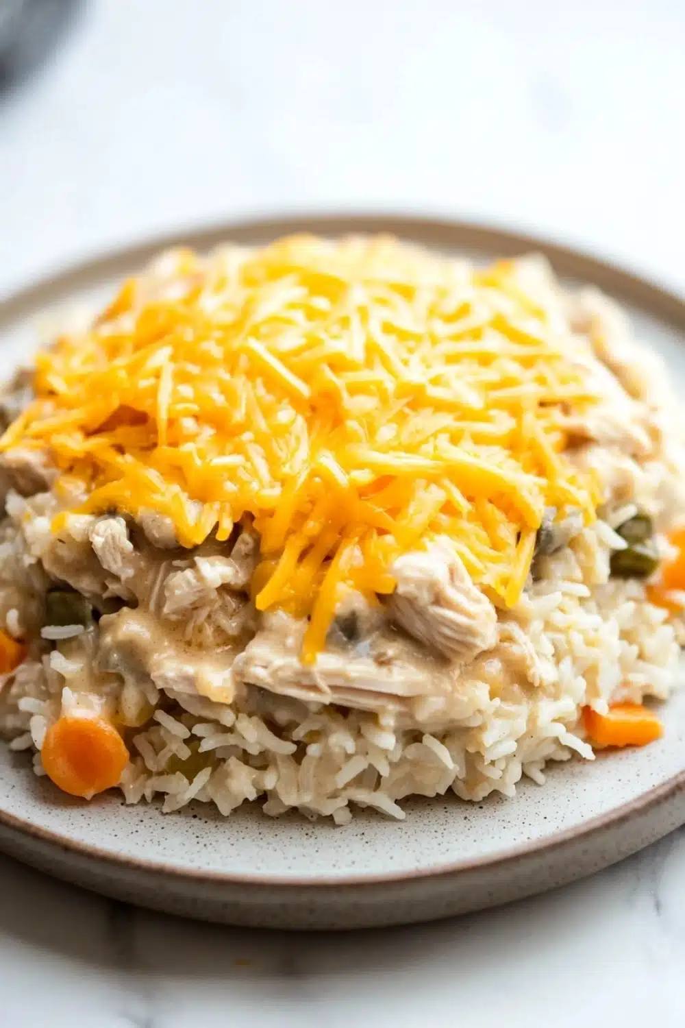 Chicken rice casserole crockpot - the image shows a plate of food with a serving of rice and chicken. the rice is white and fluffy, and the chicken is covered in a creamy sauce. on top of the rice, there is a layer of shredded cheese, which is a bright yellow color. the dish is garnished with small orange carrots and green bell peppers. the plate is sitting on a white marble countertop.