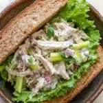 Chicken salad sandwich with yogurt - the image shows a close-up of a sandwich in a brown bowl. the sandwich is made with two slices of toasted bread and is filled with a salad. the salad is made up of lettuce, celery, onions, and shredded chicken. the chicken is white and appears to be seasoned with herbs and spices. the bowl is sitting on a wooden table with a white napkin in the background.