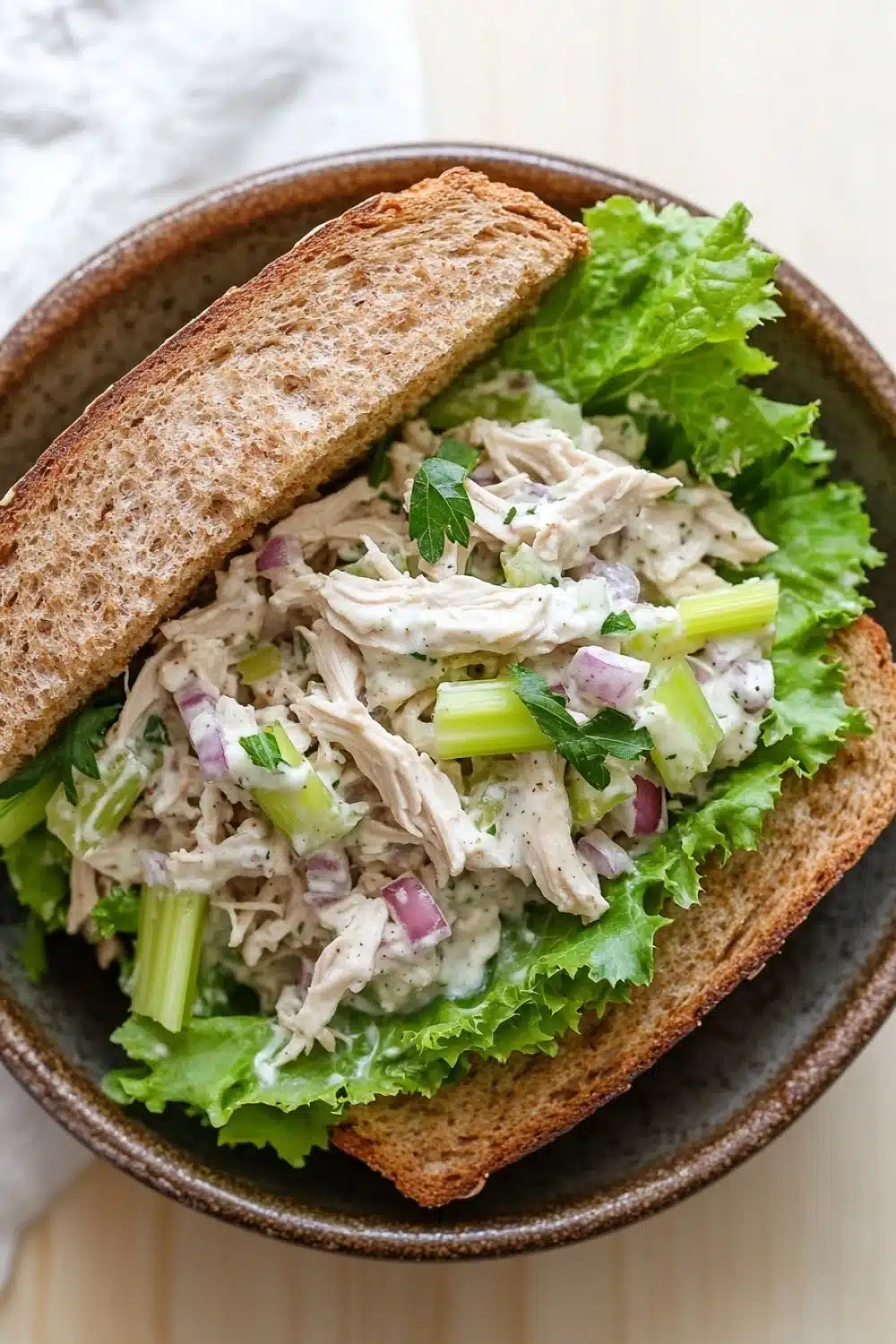Chicken salad sandwich with yogurt - the image shows a close-up of a sandwich in a brown bowl. the sandwich is made with two slices of toasted bread and is filled with a salad. the salad is made up of lettuce, celery, onions, and shredded chicken. the chicken is white and appears to be seasoned with herbs and spices. the bowl is sitting on a wooden table with a white napkin in the background.