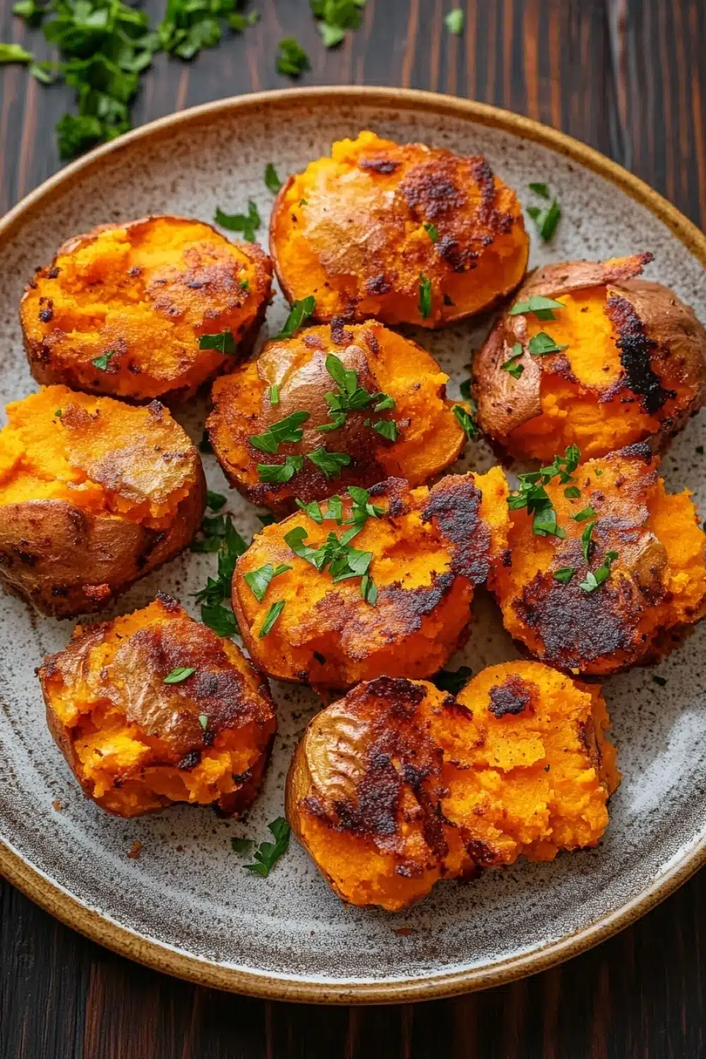 Crispy smashed sweet potatoes air fryer - the image shows a plate of roasted sweet potatoes on a wooden table. the potatoes are golden brown and appear to be seasoned with herbs and spices. they are arranged in a circular pattern on the plate, with some overlapping each other. the plate is made of light-colored wood and has a textured surface. there are a few sprigs of parsley scattered around the plate. the overall appearance of the dish is savory and appetizing.