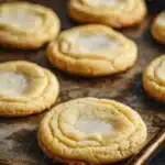 Crispy sugar cookie cut out - the image shows a tray of freshly baked cookies on a wooden table. the cookies are round and golden brown in color, with a crumbly texture on top. they are arranged in a single layer on the tray, with some overlapping each other. some of the cookies have a light dusting of powdered sugar on top, indicating that they have been freshly baked. the tray is lined with parchment paper, and the background is blurred, making the cookies the focal point of the image.