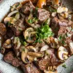 Crock pot beef mushrooms - the image is a close-up of a dish in a bowl. the dish appears to be a beef stroganoff with mushrooms and onions. the beef is cooked medium-rare and has a dark brown color. the mushrooms are sliced and caramelized, and they are arranged in a circular pattern around the beef. the onions are sliced into thin slices and are scattered throughout the dish. there is a sprig of parsley on top of the beef, adding a pop of green color to the dish and giving it a fresh and appetizing appearance. the bowl is made of a light-colored ceramic with a speckled texture. the background is a dark grey countertop.