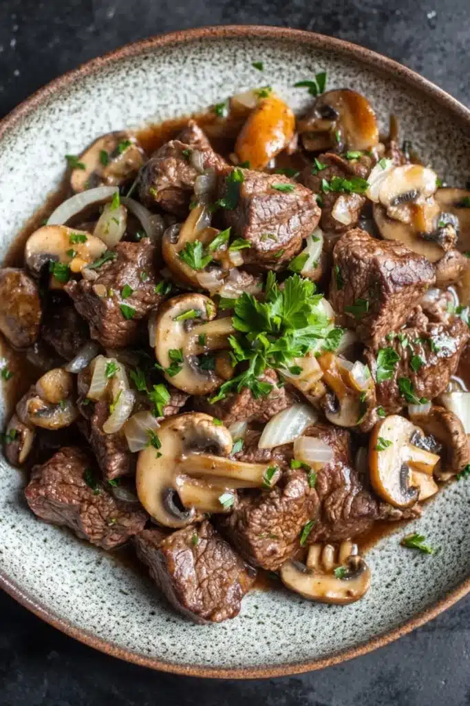 Crock pot beef mushrooms - the image is a close-up of a dish in a bowl. the dish appears to be a beef stroganoff with mushrooms and onions. the beef is cooked medium-rare and has a dark brown color. the mushrooms are sliced and caramelized, and they are arranged in a circular pattern around the beef. the onions are sliced into thin slices and are scattered throughout the dish. there is a sprig of parsley on top of the beef, adding a pop of green color to the dish and giving it a fresh and appetizing appearance. the bowl is made of a light-colored ceramic with a speckled texture. the background is a dark grey countertop.
