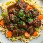Crock pot beef over noodles - the image shows a plate of beef stew with noodles. the beef is cooked in a rich, dark brown sauce and is garnished with chopped parsley. the noodles are long and thin, and they are arranged in a pile on top of the beef. there are also chunks of carrots and celery scattered throughout the dish. the plate is white with a blue rim and is sitting on a white marble countertop.