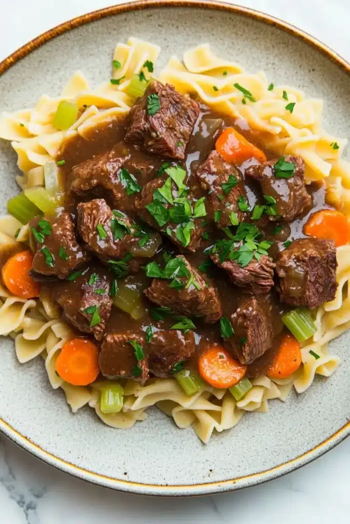 Crock pot beef over noodles - the image shows a plate of beef stew with noodles. the beef is cooked in a rich, dark brown sauce and is garnished with chopped parsley. the noodles are long and thin, and they are arranged in a pile on top of the beef. there are also chunks of carrots and celery scattered throughout the dish. the plate is white with a blue rim and is sitting on a white marble countertop.
