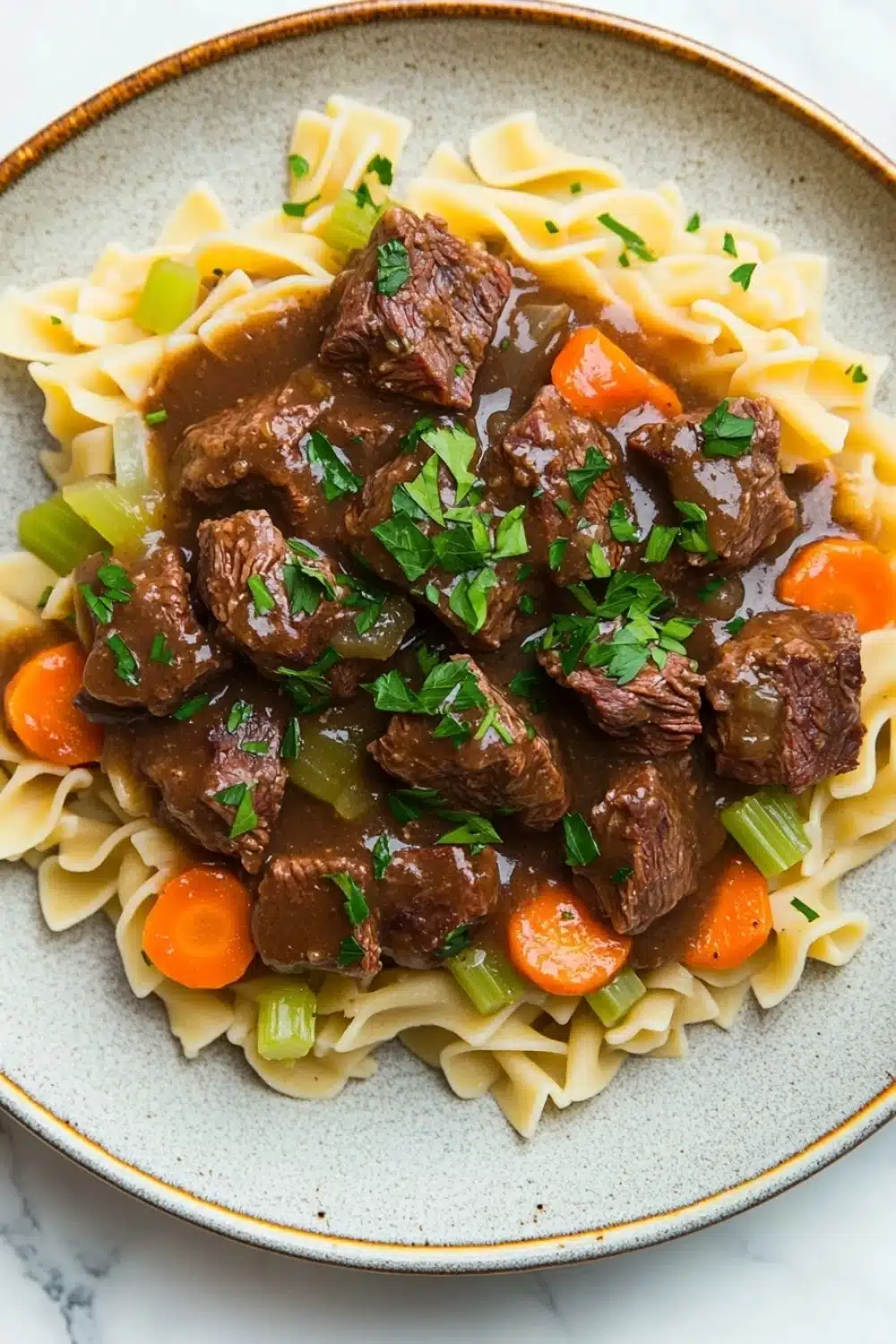 Crock pot beef over noodles - the image shows a plate of beef stew with noodles. the beef is cooked in a rich, dark brown sauce and is garnished with chopped parsley. the noodles are long and thin, and they are arranged in a pile on top of the beef. there are also chunks of carrots and celery scattered throughout the dish. the plate is white with a blue rim and is sitting on a white marble countertop.