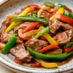 Crock pot beef pepper steak - the image is a close-up of a plate of food. the plate is white with a speckled pattern and is placed on a wooden table. the food appears to be a stir-fry dish with chunks of beef and colorful bell peppers. the beef is cooked medium-rare and the peppers are bright red, yellow, and green. the peppers are arranged in a pile on top of the beef, with some overlapping each other. the sauce is a dark brown color and is drizzled over the top. the dish looks appetizing and ready to eat.