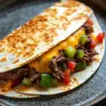 Crock pot beef quesadilla - the image shows a plate with a quesadilla on it. the plate is made of dark grey ceramic with a speckled pattern. on top of the quesads, there is a layer of shredded beef, diced red and green bell peppers, and a dollop of melted cheese. the beef appears to be cooked medium-rare and is cut into small pieces. the cheese is a bright yellow color and is melted and bubbly. the peppers are bright red and appear to be seasoned with herbs and spices. the dish looks appetizing and ready to eat.