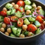 Cucumber tomato salad garbanzo - the image is a close-up of a colorful salad in a black bowl. the salad is made up of diced tomatoes, cucumbers, chickpeas, and red onions. the tomatoes are bright red and appear to be ripe and juicy. the cucumbers are cut into small cubes and are scattered throughout the salad. the chickpea is light brown and is scattered throughout. there are also some green herbs, possibly parsley, adding a pop of color to the dish. the bowl is sitting on a dark blue surface with a few sprigs of parsley scattered around it.