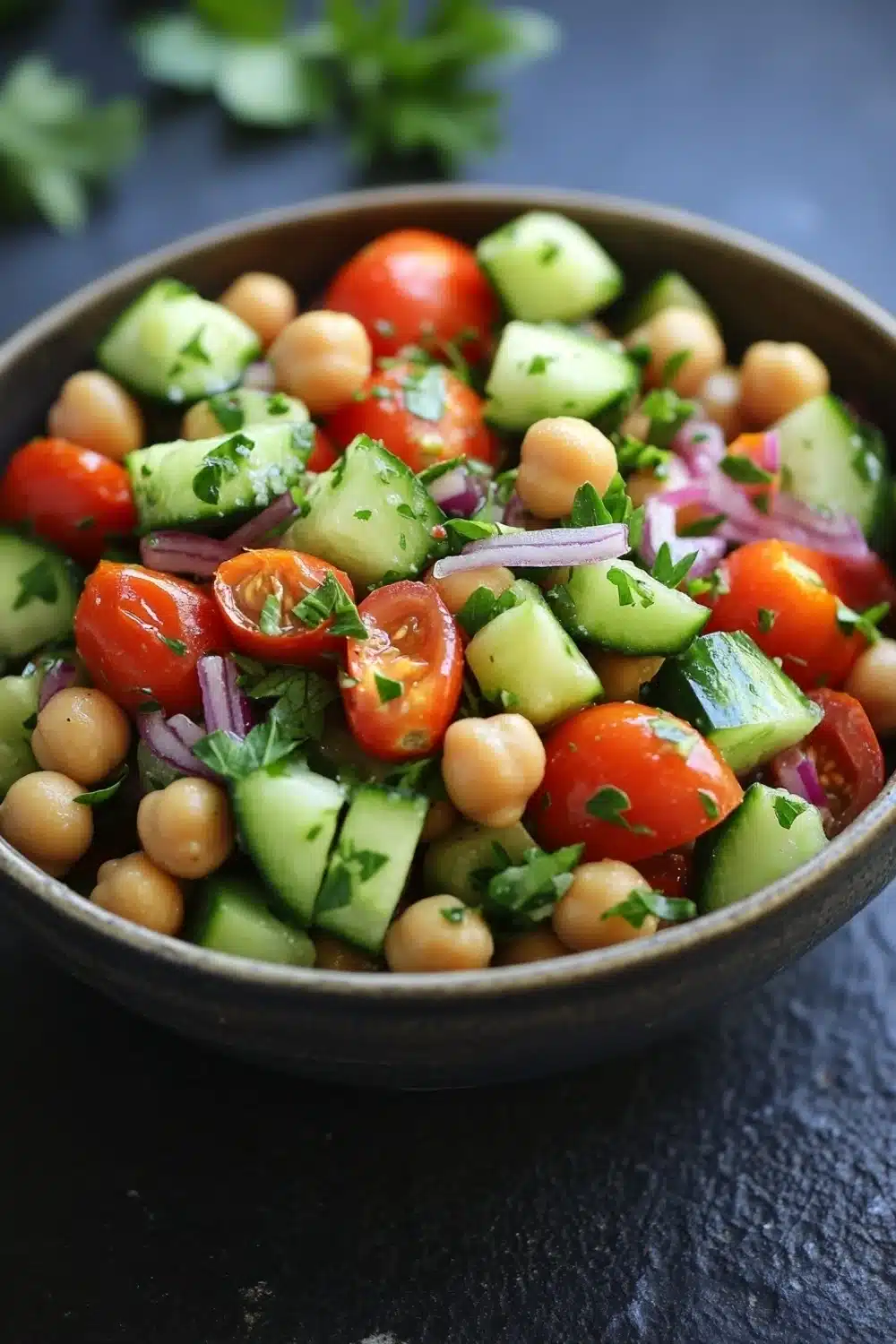 Cucumber tomato salad garbanzo - the image is a close-up of a colorful salad in a black bowl. the salad is made up of diced tomatoes, cucumbers, chickpeas, and red onions. the tomatoes are bright red and appear to be ripe and juicy. the cucumbers are cut into small cubes and are scattered throughout the salad. the chickpea is light brown and is scattered throughout. there are also some green herbs, possibly parsley, adding a pop of color to the dish. the bowl is sitting on a dark blue surface with a few sprigs of parsley scattered around it.