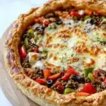 Deep dish pizza pot pie - the image is of a freshly baked pizza on a wooden cutting board. the pizza has a golden brown crust and is topped with melted cheese, black olives, red bell peppers, and ground beef. the toppings are arranged in a circular pattern around the edges of the crust. the background is a white marble countertop.
