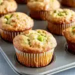 Green apple muffin - the image shows a muffin tin filled with freshly baked muffins. the muffins are golden brown in color and have a crumbly texture on top. they are arranged in a single layer on the tin, with some overlapping each other. some of the muffins have a bite taken out of one of them, revealing the green filling inside. the tin is placed on a white marble countertop.