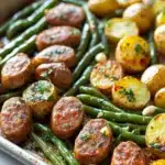 Green bean sausage sheet pan dinner - the image shows a baking tray filled with roasted potatoes and green beans. the potatoes are golden brown and appear to be seasoned with herbs and spices. the green beans are long and slender, and they are arranged in a single layer on the tray. the tray is lined with parchment paper, and there are small pieces of garlic scattered throughout the dish. the background is blurred, but it appears to be a kitchen countertop.