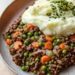 Ground beef dinner for kids - the image shows a plate of food with a serving of mashed potatoes on top. the mashed potatoes are white and fluffy, and the dish appears to be a mixture of ground beef, carrots, peas, onions, and other vegetables. the plate is brown and has a speckled pattern around the edges. the food is garnished with a sprig of fresh herbs. the background is a wooden table.