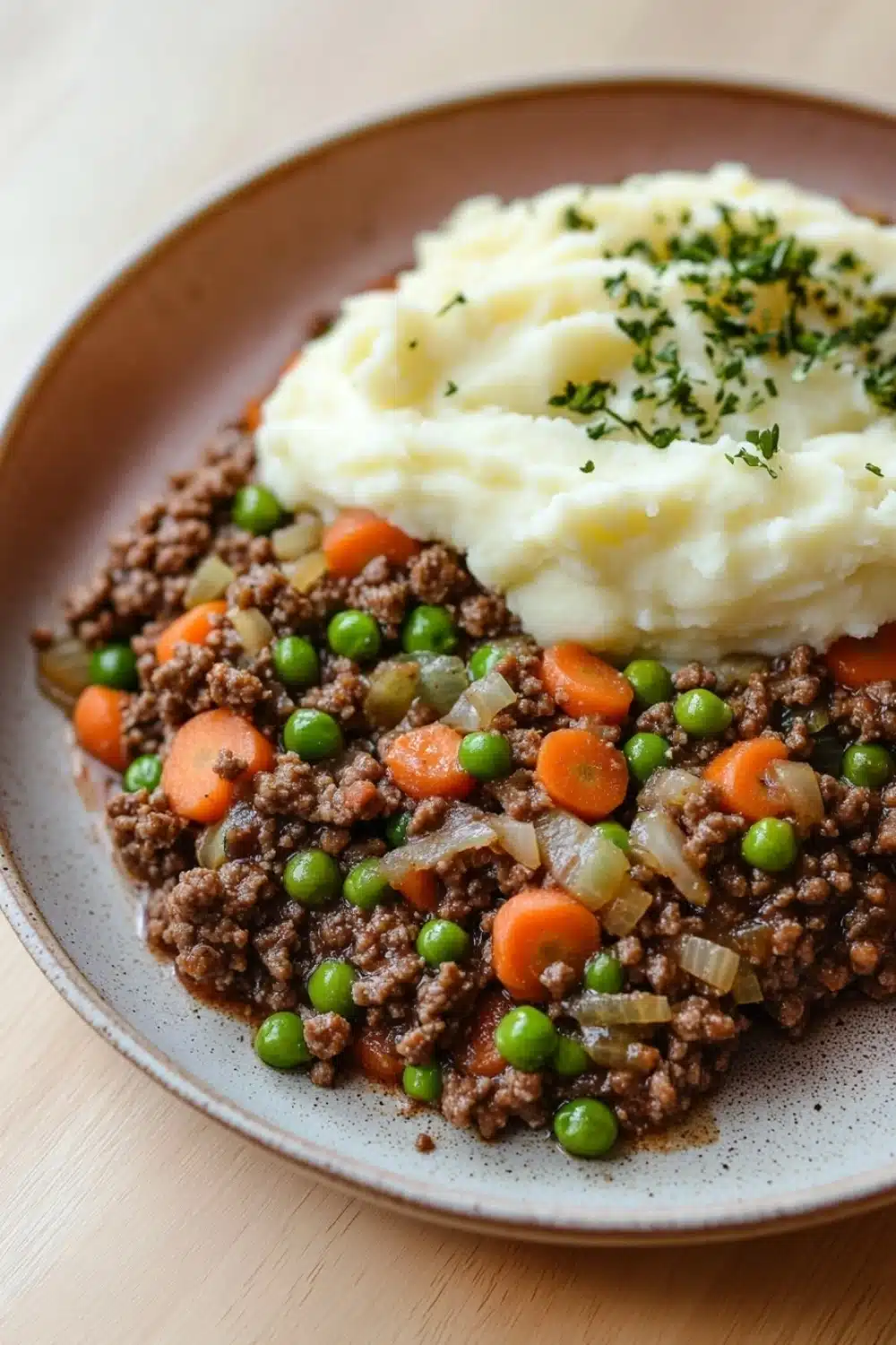 Ground beef dinner for kids - the image shows a plate of food with a serving of mashed potatoes on top. the mashed potatoes are white and fluffy, and the dish appears to be a mixture of ground beef, carrots, peas, onions, and other vegetables. the plate is brown and has a speckled pattern around the edges. the food is garnished with a sprig of fresh herbs. the background is a wooden table.