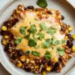 High protein beef enchilada skillet - the image shows a plate of food on a white marble countertop. the plate is round and has a light brown color. on top of the plate, there is a layer of ground beef, black beans, corn kernels, and rice. the beef appears to be cooked and has melted cheese on top. there are also a few sprigs of cilantro scattered around the plate. the overall presentation of the dish is colorful and appetizing.