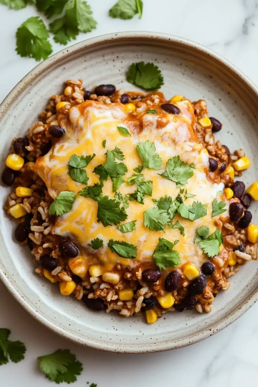 High protein beef enchilada skillet - the image shows a plate of food on a white marble countertop. the plate is round and has a light brown color. on top of the plate, there is a layer of ground beef, black beans, corn kernels, and rice. the beef appears to be cooked and has melted cheese on top. there are also a few sprigs of cilantro scattered around the plate. the overall presentation of the dish is colorful and appetizing.