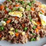 High protein korean beef fried rice - the image is a close-up of a plate of fried rice. the rice is brown and appears to be seasoned with minced meat and vegetables. there are small pieces of orange and green peas scattered throughout the dish. on top of the rice, there are two hard-boiled eggs. the plate is white with a speckled pattern. the background is a light beige color.