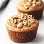 High protein vegan breakfast muffin - the image shows two freshly baked muffins on a white baking tray. the muffins are golden brown in color and have a crumbly texture. they are topped with chopped walnuts, giving them a crunchy appearance. the tray is lined with parchment paper and there is a blurred background of other muffins.