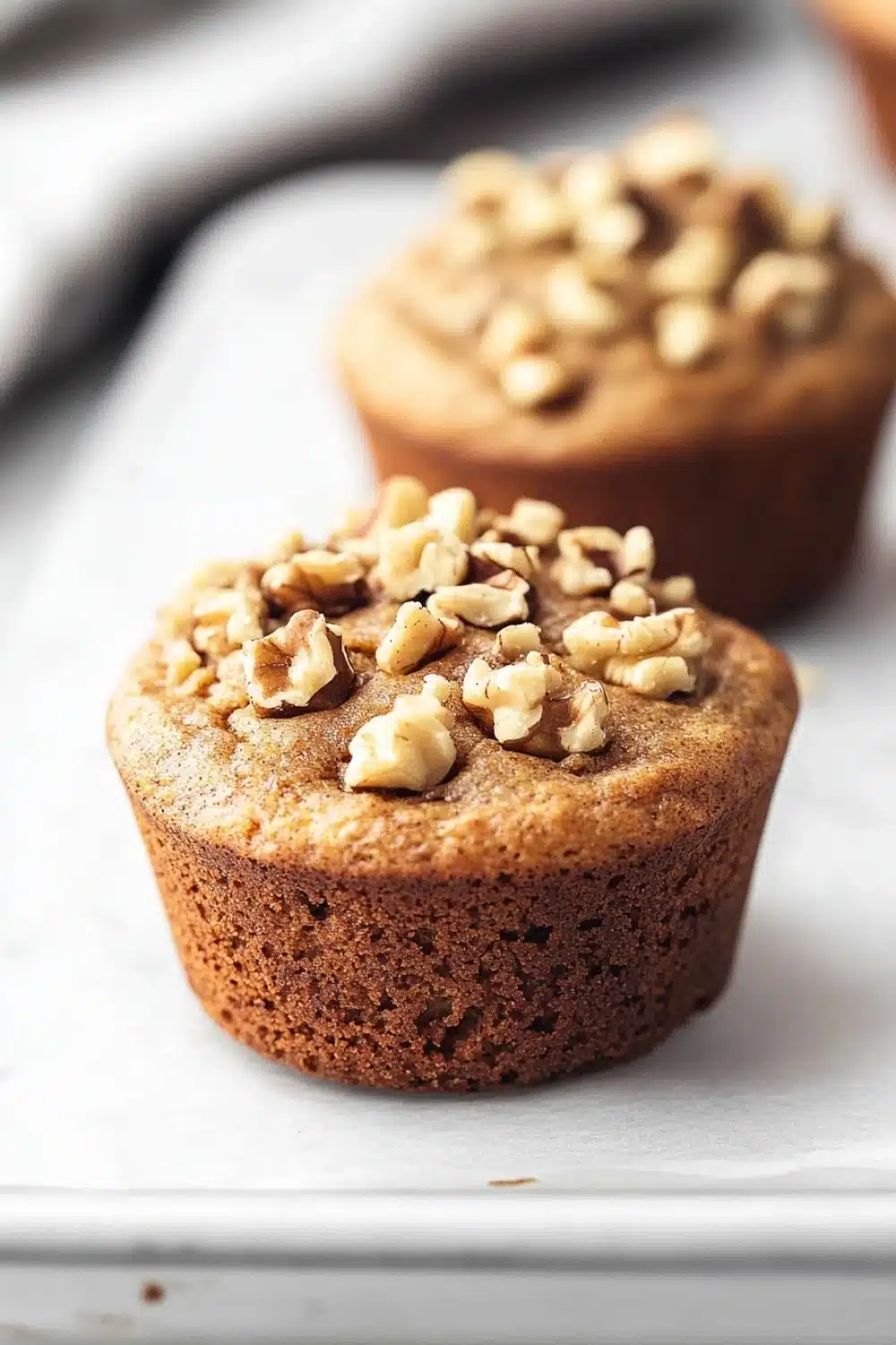 High protein vegan breakfast muffin - the image shows two freshly baked muffins on a white baking tray. the muffins are golden brown in color and have a crumbly texture. they are topped with chopped walnuts, giving them a crunchy appearance. the tray is lined with parchment paper and there is a blurred background of other muffins.