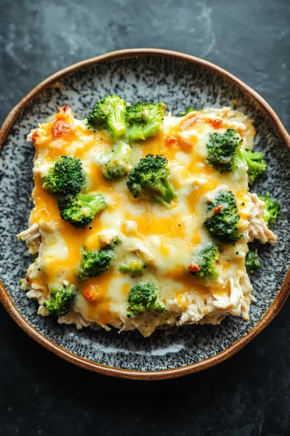 Keto chicken broccoli casserole - the image is a close-up of a dish in a brown ceramic bowl. the dish appears to be a casserole or lasagna, with a layer of melted cheese on top. the cheese is yellow and bubbly, and there are small pieces of broccoli scattered throughout the dish. the broccoli is bright green and looks fresh and healthy. the bowl is sitting on a dark grey countertop.