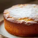 Lemon cake oil - the image is a close-up of a round cake on a white plate. the cake appears to be freshly baked and has a golden brown color. it is dusted with powdered sugar, giving it a shiny appearance. the plate is sitting on a wooden table, and the background is blurred, making the cake the focal point of the image.