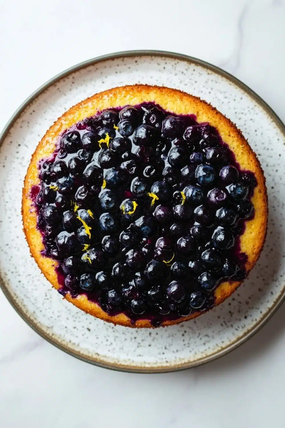 Lemon cake with blueberry pie filling - the image is of a round cake on a white plate with a speckled pattern. the cake appears to be a blueberry cake with a golden brown crust and a layer of blueberries on top. the blueberries are arranged in a circular pattern around the edges of the cake, with some of them overlapping each other. there are also some orange zest scattered throughout the cake. the plate is sitting on a marble countertop.