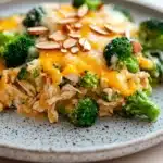 Macro chicken broccoli casserole - the image is a close-up of a plate of food. the plate is white with a speckled pattern and is placed on a wooden table. on top of the plate, there is a serving of broccoli casserole. the broccoli is cut into small pieces and is covered in a layer of melted cheese. there are also some sliced almonds scattered on top. the dish appears to be freshly cooked and ready to eat.