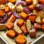 Maple roasted root vegetables - the image shows a baking tray filled with roasted sweet potatoes and red onions. the potatoes are golden brown and appear to be seasoned with herbs and spices. the carrots are cut into small cubes and are arranged in a single layer on the tray. the onions are sliced into thin strips and are scattered throughout the tray, adding a pop of color to the dish. the tray is lined with parchment paper and is sitting on a wooden table.