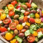 Mediterranean marinated vegetable salad - the image is a close-up of a colorful salad in a bowl. the salad is made up of various types of vegetables, including cherry tomatoes, cucumbers, olives, red onions, and feta cheese. the tomatoes are bright red and appear to be ripe and juicy. the cucumbers are cut into small cubes and are scattered throughout the salad. the olives are dark red and look ripe and ready to eat. the feta is crumbled and adds a creamy texture to the dish. the bowl is made of a light-colored material and is sitting on a white marble countertop.