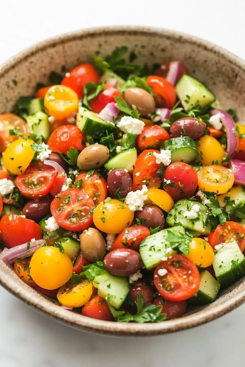 Mediterranean marinated vegetable salad - the image is a close-up of a colorful salad in a bowl. the salad is made up of various types of vegetables, including cherry tomatoes, cucumbers, olives, red onions, and feta cheese. the tomatoes are bright red and appear to be ripe and juicy. the cucumbers are cut into small cubes and are scattered throughout the salad. the olives are dark red and look ripe and ready to eat. the feta is crumbled and adds a creamy texture to the dish. the bowl is made of a light-colored material and is sitting on a white marble countertop.